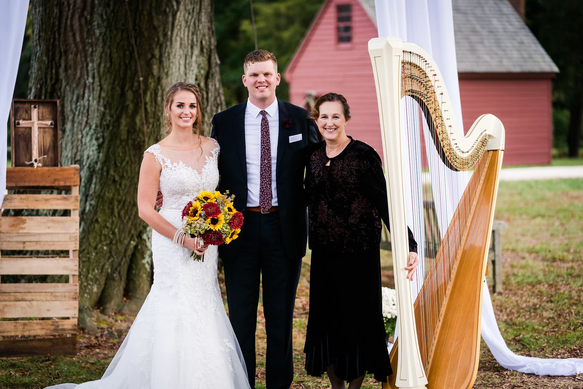 Bride, groom, and woman posing outdoors near a harp.