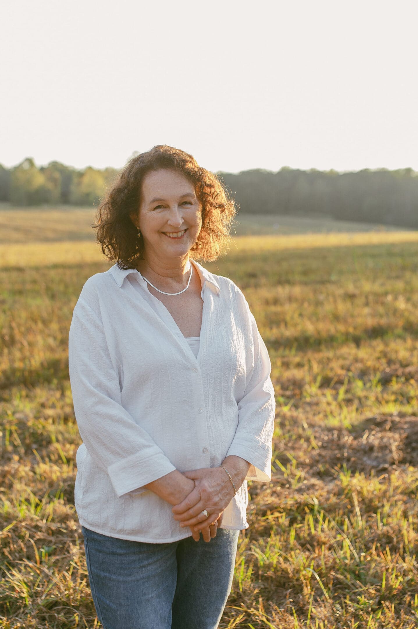 Woman in white shirt standing in a sunlit field, smiling.