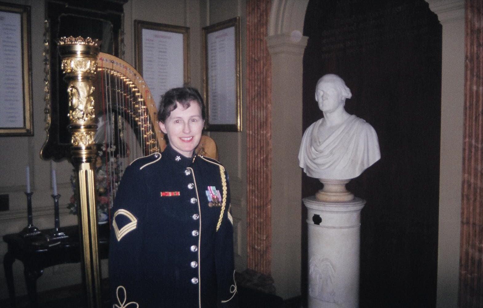 Person in formal military dress standing indoors near a classical bust.